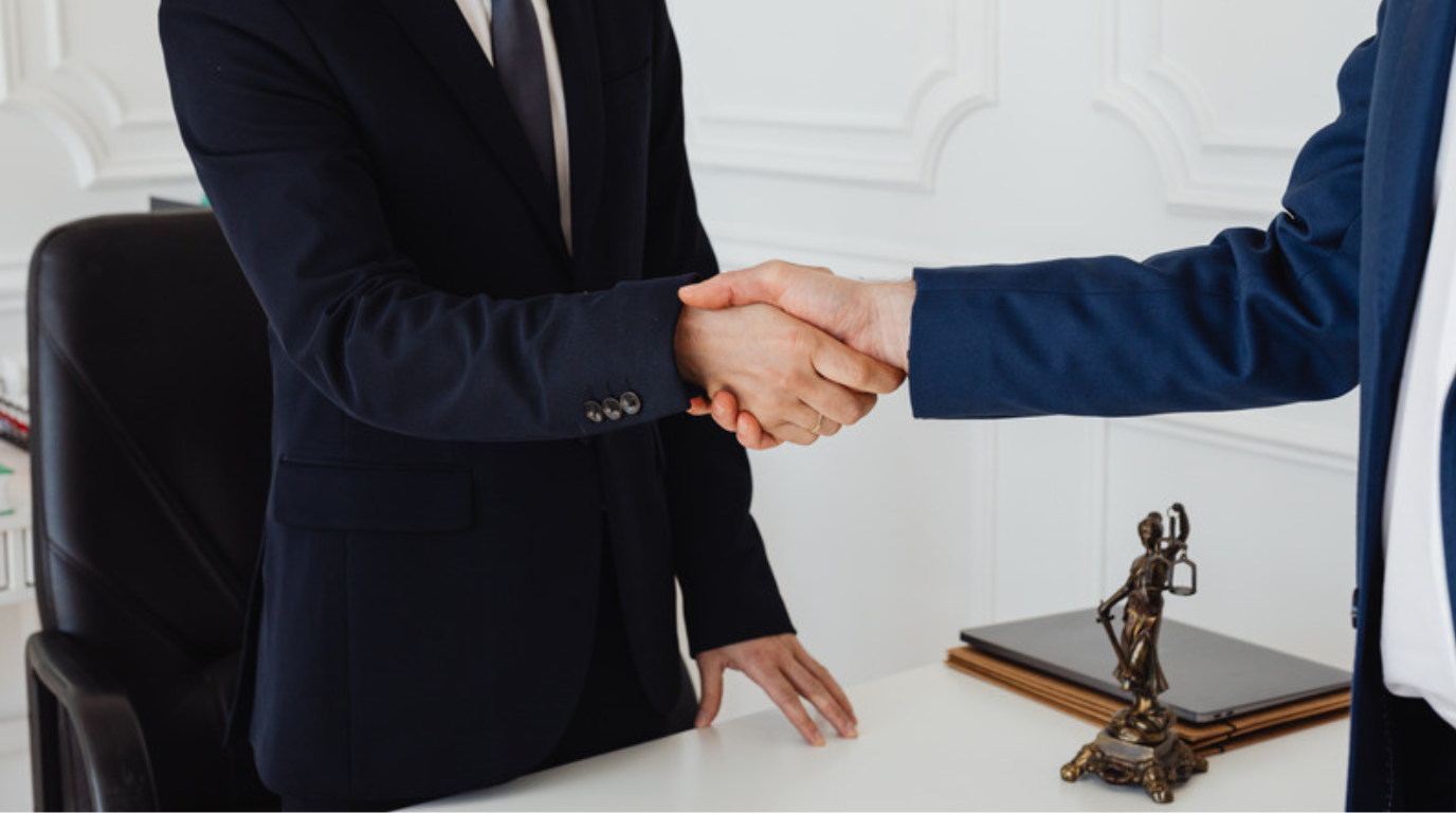 Two people in suits shake hands across a desk in an office, with a stack of folders and a small statue of Lady Justice visible—an ideal scene for a Personal Injury Attorney in Midway, Utah.